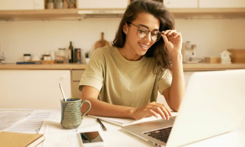 Charming Hispanic student girl in stylish glasses doing homework at kitchen table, learning distantly from home, using generic laptop. Attractive young female freelancer working on portable computer